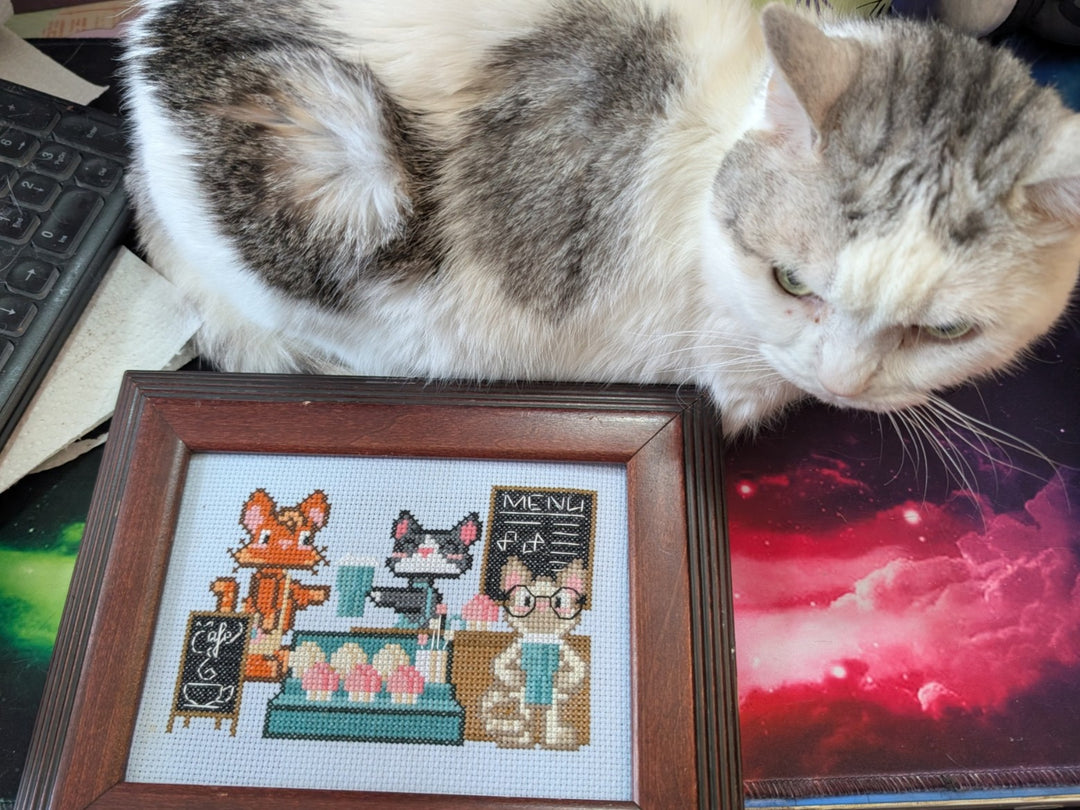 A gray and white cat lies on a desk next to a framed pixel-art cross-stitch design of a cat cafe scene.