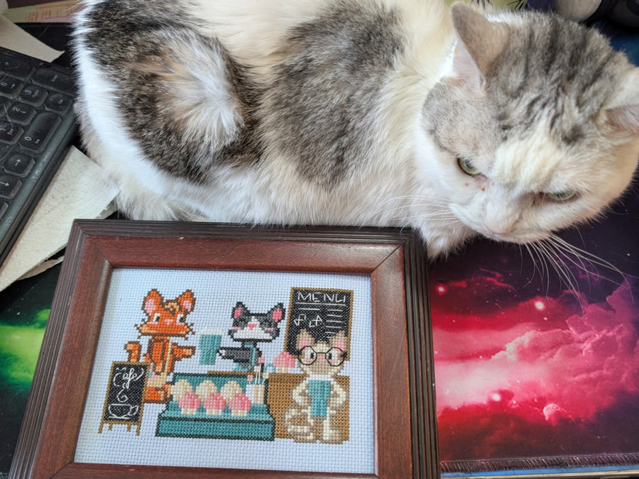 A gray and white cat lies on a desk next to a framed pixel-art cross-stitch design of a cat cafe scene.