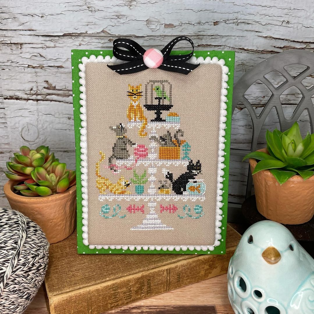 Cross-stitched picture of cats on a stand with decorative elements, framed in green, on a wooden surface.
