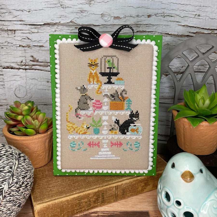Cross-stitched picture of cats on a stand with decorative elements, framed in green, on a wooden surface.