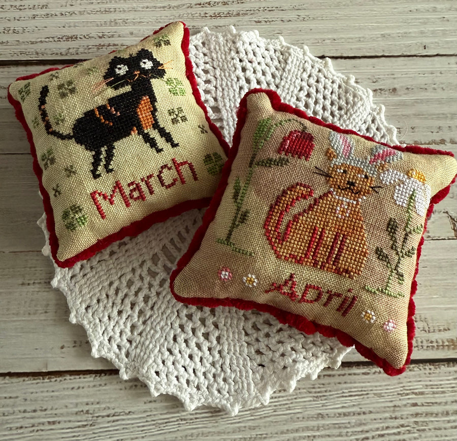 Two needlecraft pillows, one labeled 'March' with a black cat design and the other labeled 'April' with an orange cat design, placed on a white doily.