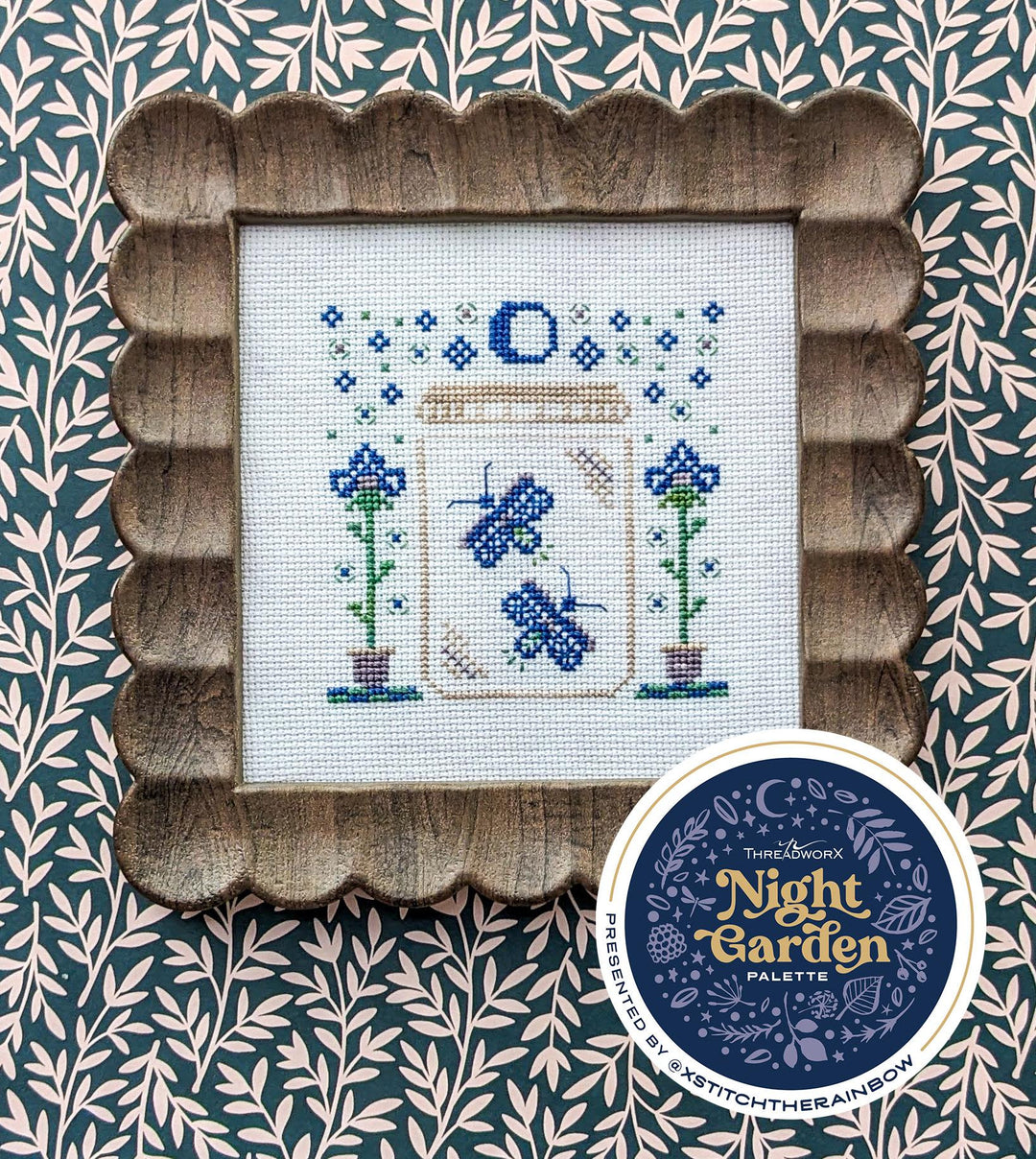 Cross-stitched design of a garden scene with a wooden frame on a patterned background, featuring the 'Night Garden Palette' brand.
