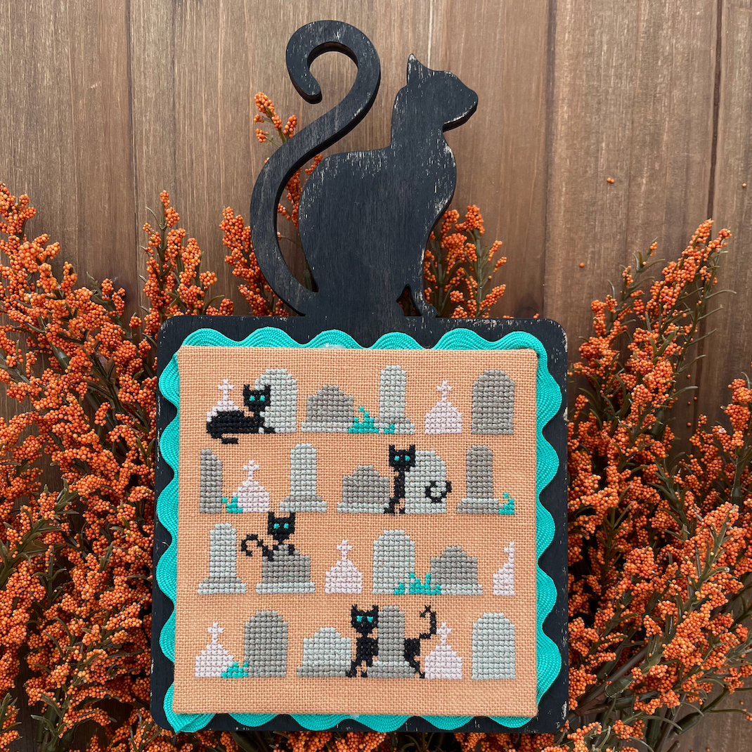 Cross-stitched design of a cat and tombs on a wooden background with decorative plants.