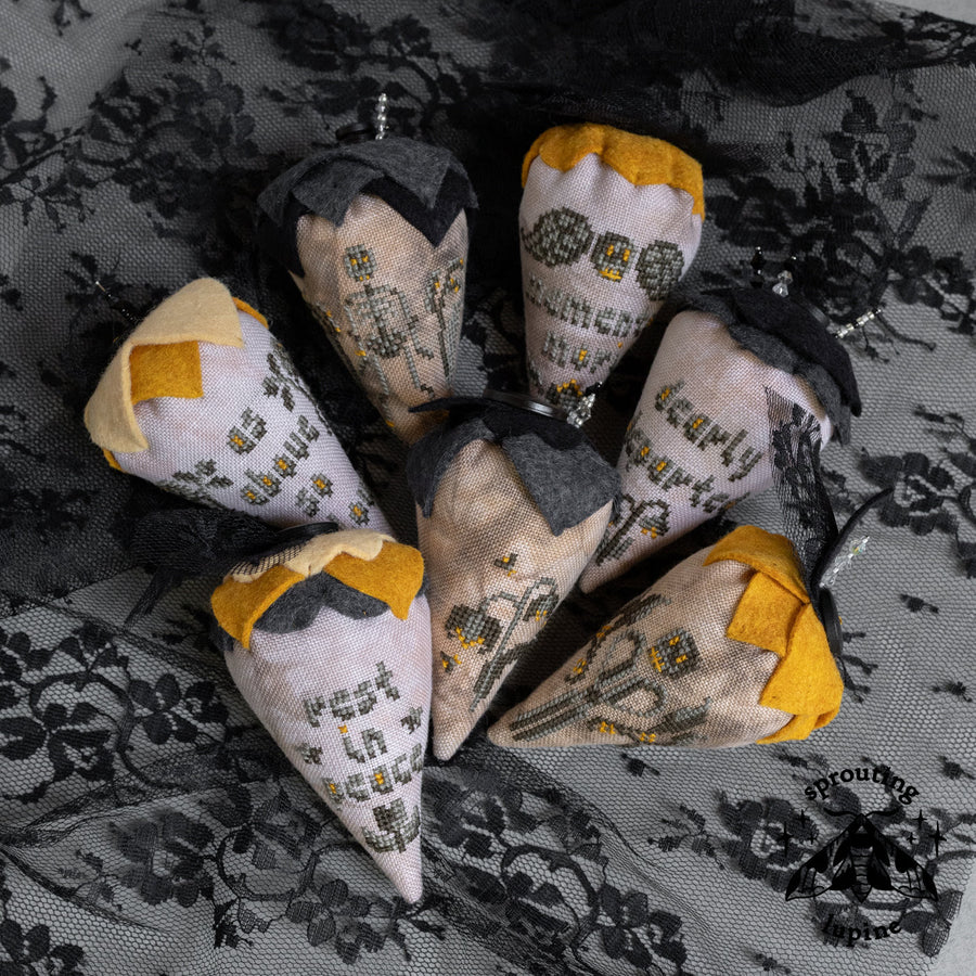 seven handmade fabric cone-shaped ornaments with embroidered floral and text designs in muted yellow, gray, black, and off-white, arranged on a black lace fabric background.