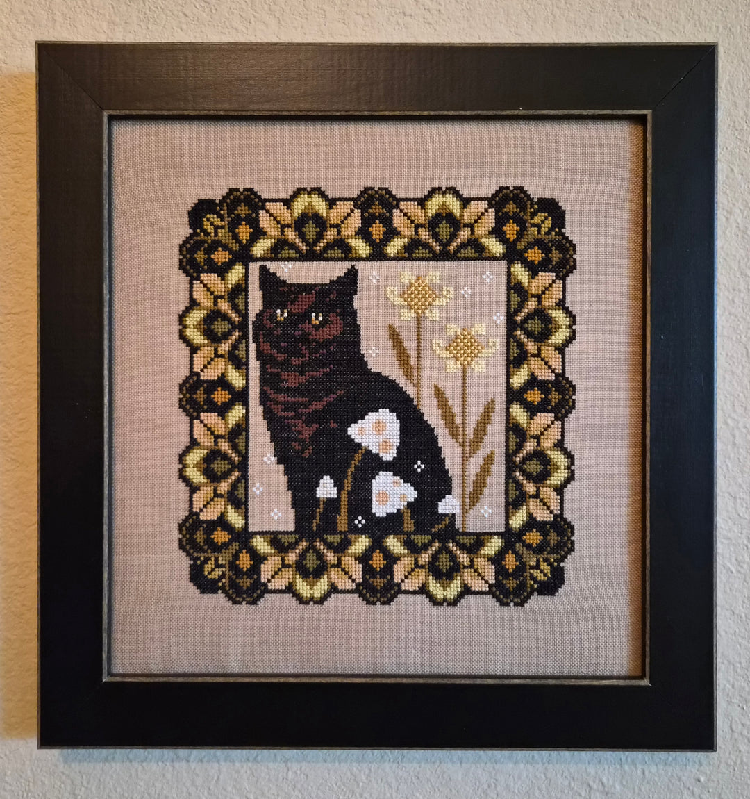 Framed cross-stitched picture of a black cat with flowers on beige fabric.