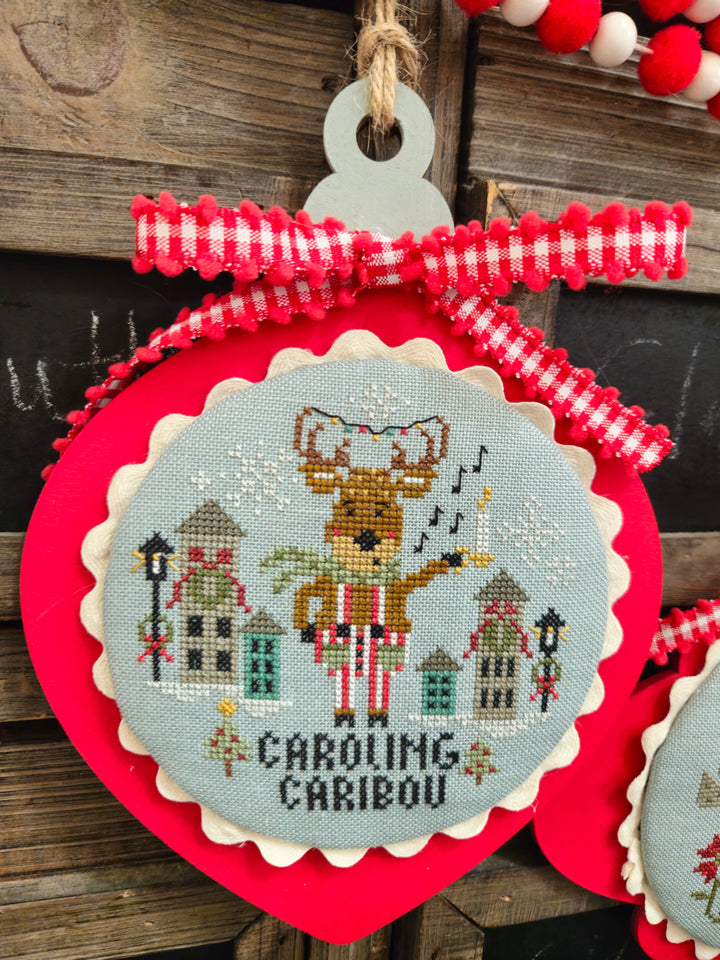 Decorative ornament with cross-stitched design of a reindeer and text 'Caroling Caribou' on a wooden background.
