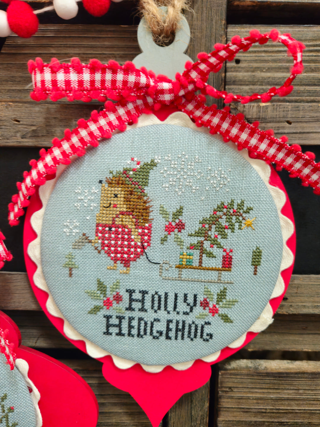 Decorative ornament with cross-stitched design of a hedgehog and text 'Holly Hedgehog' on a wooden background.