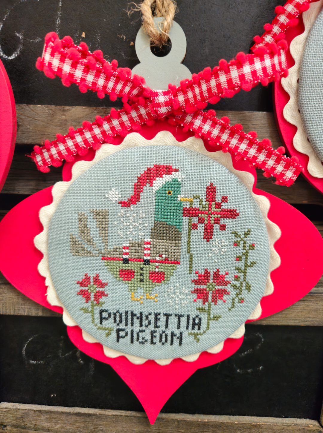Decorative ornament with cross-stitched design of a pigeon and poinsettias, tied with red and white checkered ribbon.