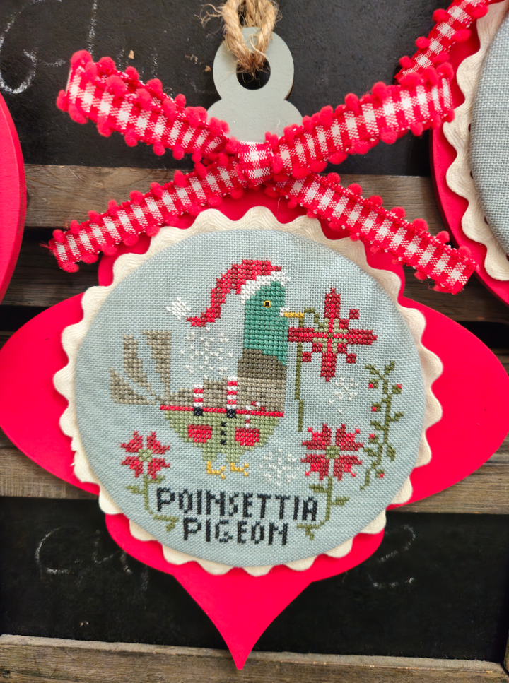 Decorative ornament with cross-stitched design of a pigeon and poinsettias, tied with red and white checkered ribbon.