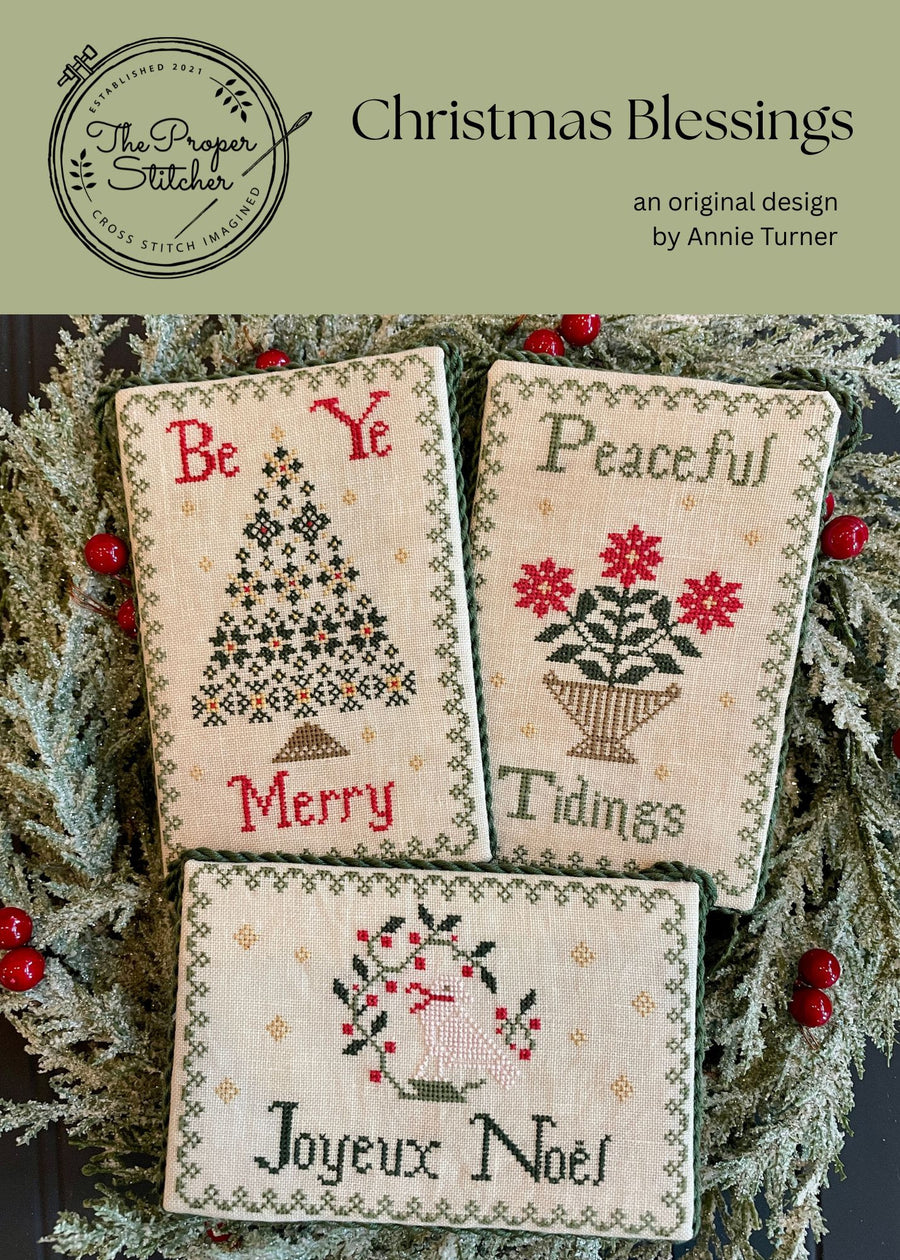 Three cross-stitched cards with Christmas-themed designs and text, framed by greenery and berries.
