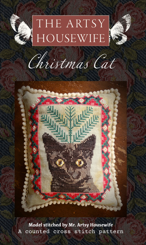 Cross-stitched cat design on a pillow with decorative border, titled 'Christmas Cat' from 'The Artsy Housewife'.
