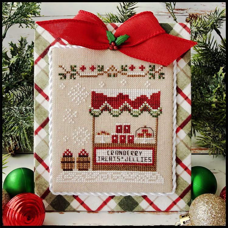 A framed cross-stitch sampler featuring a holiday storefront design with the text 'CRANBERRY TREATS JELLIES,' surrounded by a red ribbon and green pine branches with ornaments.