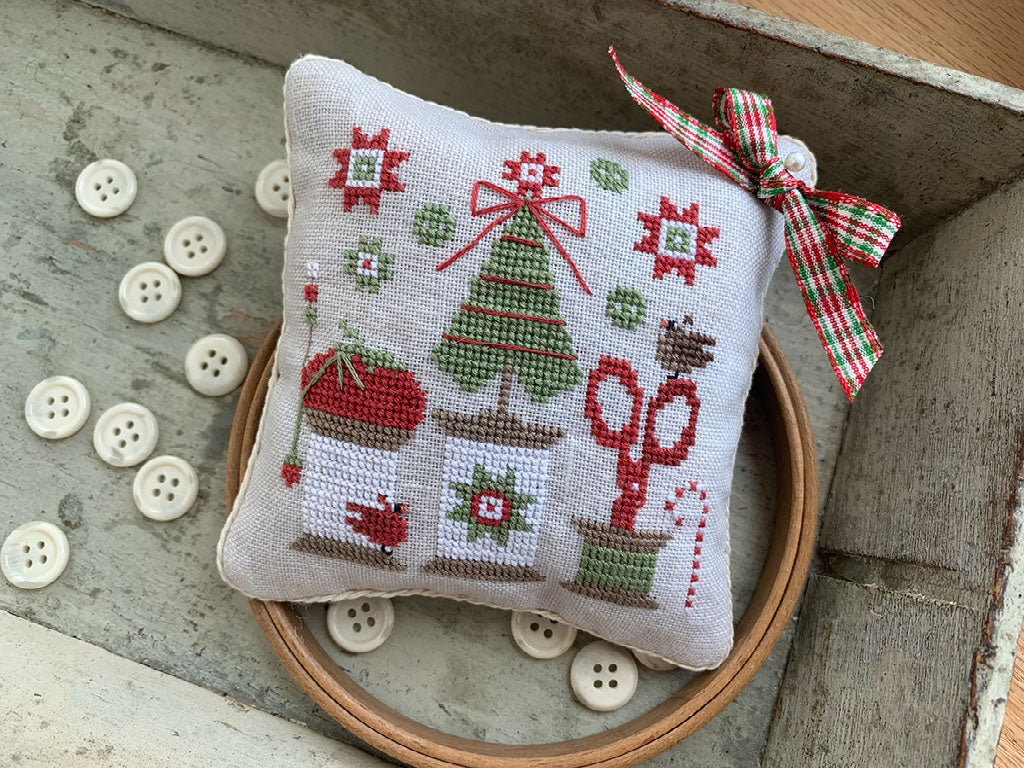 A small rectangular cross-stitch pillow with a Christmas theme, featuring a green Christmas tree, red bow, scissors, and snowman on a light beige fabric background. The pillow is decorated with a red and green plaid ribbon and is displayed inside a wooden hoop with buttons on a rustic gray surface.