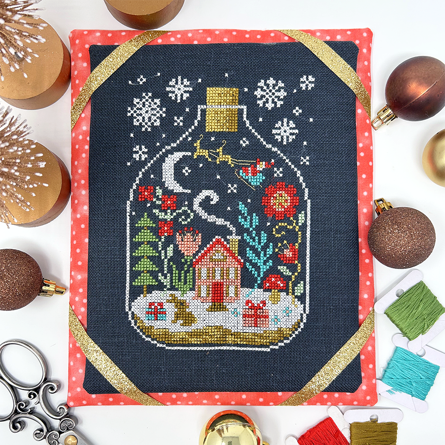 Cross-stitched design of a house in a bottle with Christmas elements, surrounded by festive decorations.
