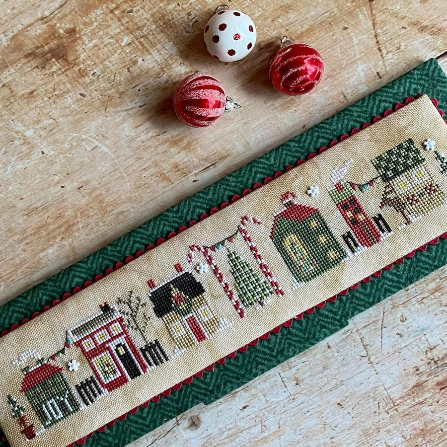 Cross-stitched panel with festive design on a wooden surface with Christmas ornaments.