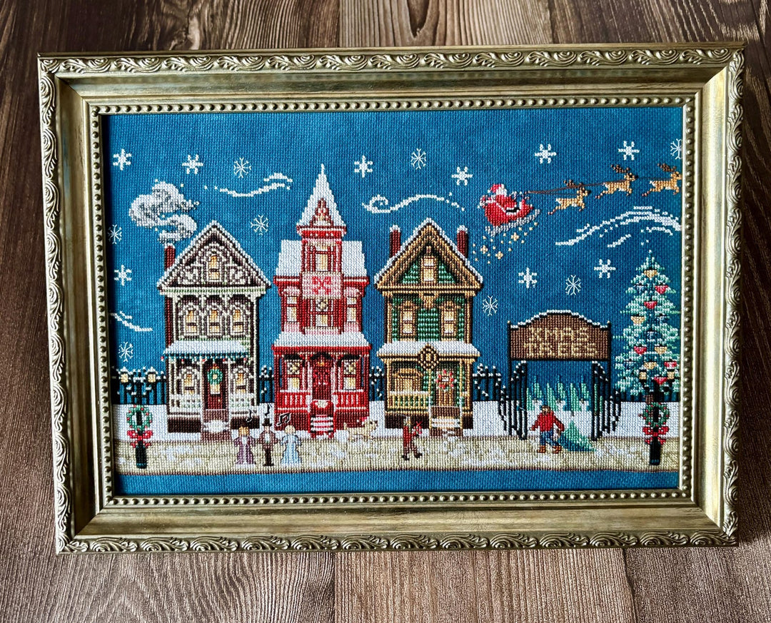 A framed Christmas cross-stitch artwork depicting three snow-covered houses, Santa Claus in his sleigh, snowflakes, and a Christmas tree, set in an ornate gold frame on a wooden surface.