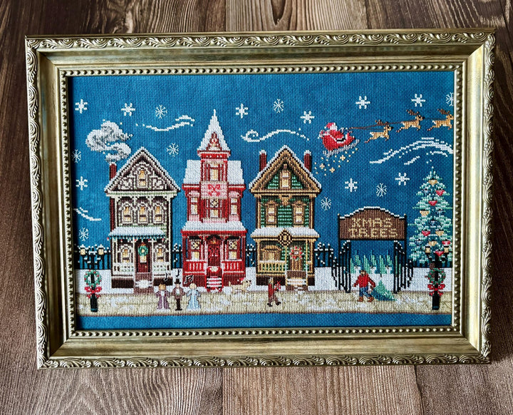 A framed Christmas cross-stitch artwork depicting three snow-covered houses, Santa Claus in his sleigh, snowflakes, and a Christmas tree, set in an ornate gold frame on a wooden surface.