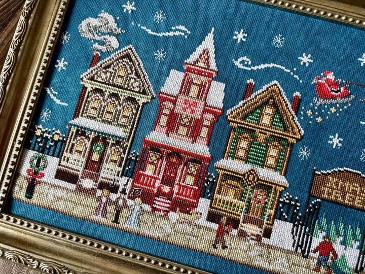 A framed cross-stitch artwork depicting a snowy Christmas village scene with three decorated houses, snowflakes, Santa in a sleigh, and small figures on a blue background.