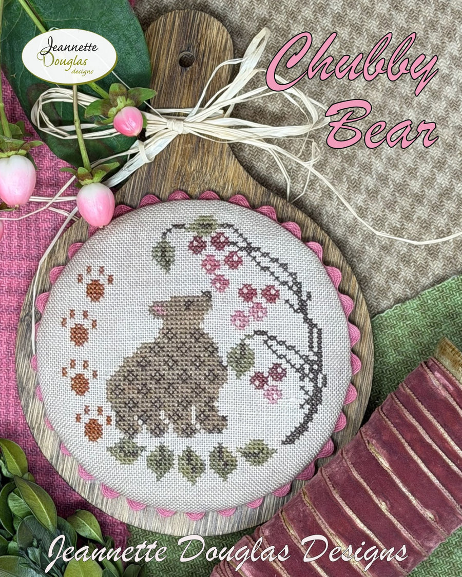 Cross-stitched round design featuring a bear surrounded by flowers and leaves, with 'Jeannette Douglas Designs' branding.