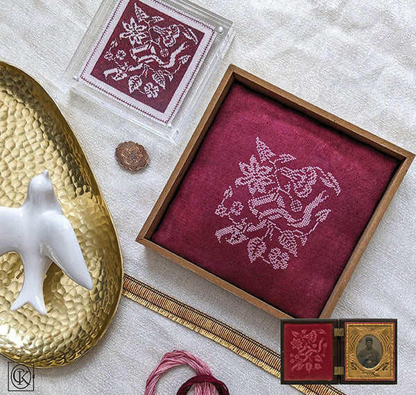 Cross stitch pattern with embroidery on fabric, surrounded by decorative items on a textured surface.
