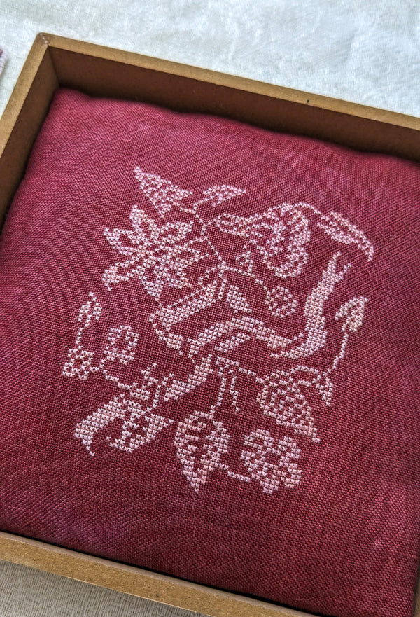Embroidered design on a red fabric inside a wooden frame.