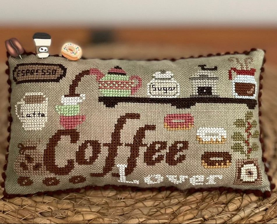 A small rectangular cross-stitch pillow with a coffee-themed design, featuring the words 'Coffee Lover' and illustrations of coffee cups, mugs, and a coffee train, displayed on a woven mat.