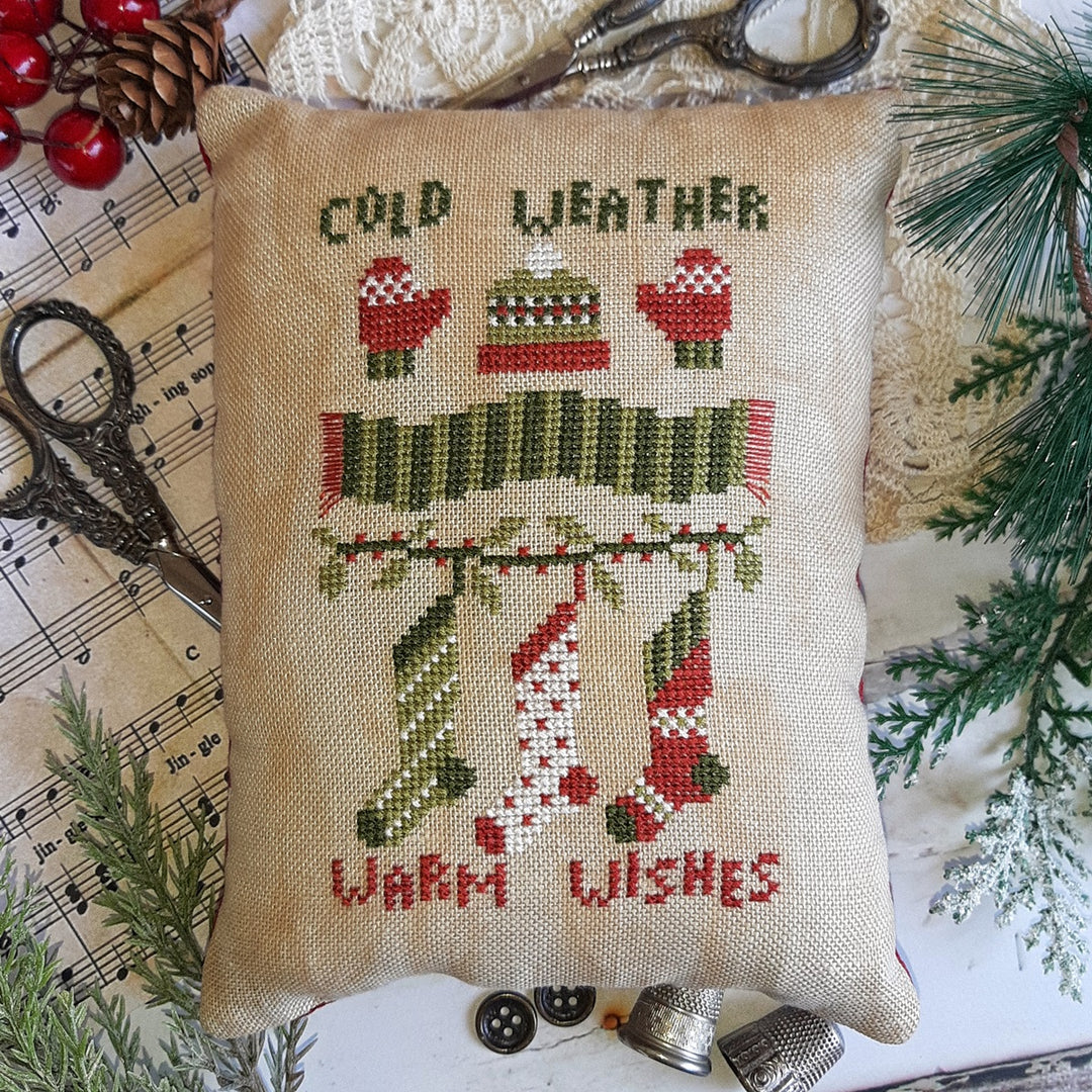 A beige throw pillow with the text 'COLD WEATHER' and 'WARM WISHES' embroidered in red and green. The pillow is placed on a white surface with pine branches in the background and a pair of scissors to the left.