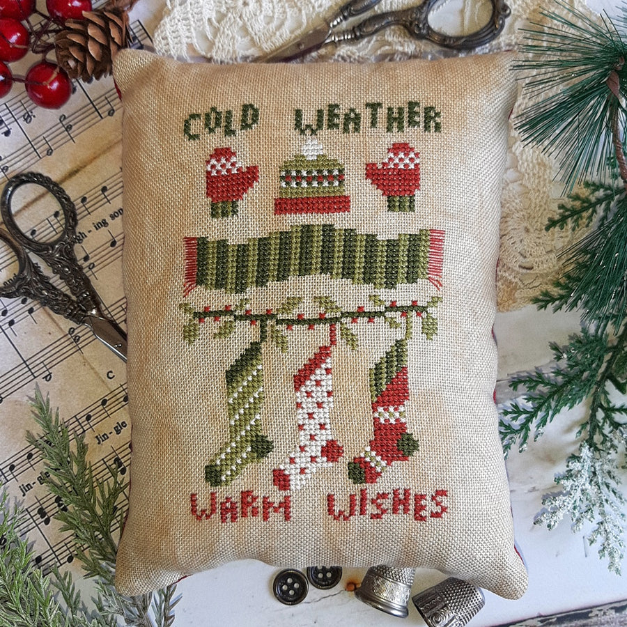 A beige throw pillow with the text 'COLD WEATHER' and 'WARM WISHES' embroidered in red and green. The pillow is placed on a white surface with pine branches in the background and a pair of scissors to the left.