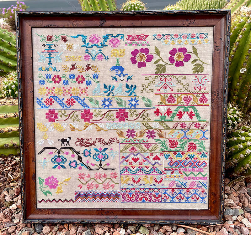 A framed cross-stitch kit pattern titled 'Colors of Mexico' with various colorful floral and geometric designs on a beige background, displayed in a garden setting.