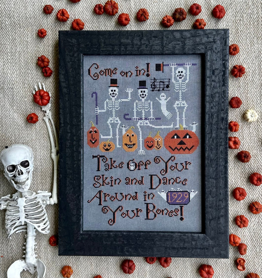 Framed Halloween-themed cross-stitch art with skeletons and pumpkins on a textured fabric background.