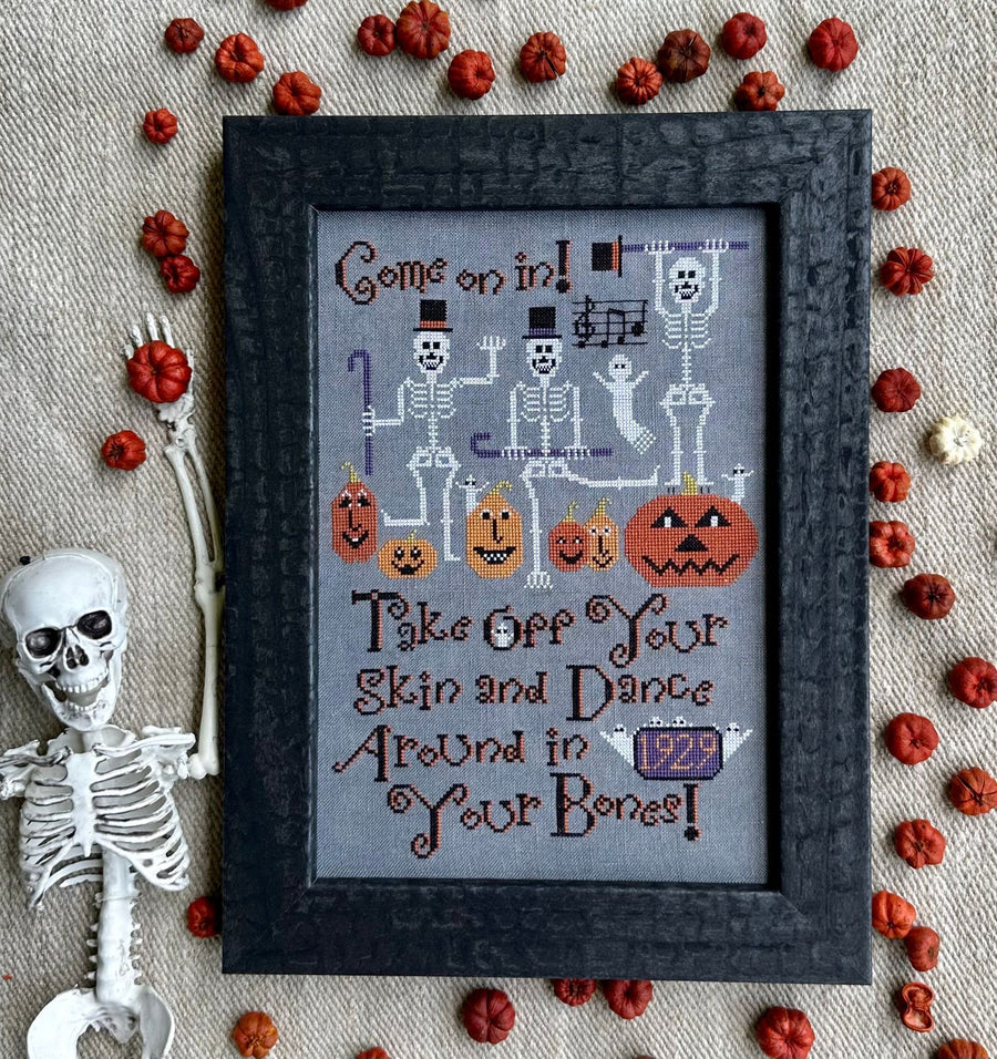 Framed Halloween-themed cross-stitch art with skeletons and pumpkins on a textured fabric background.