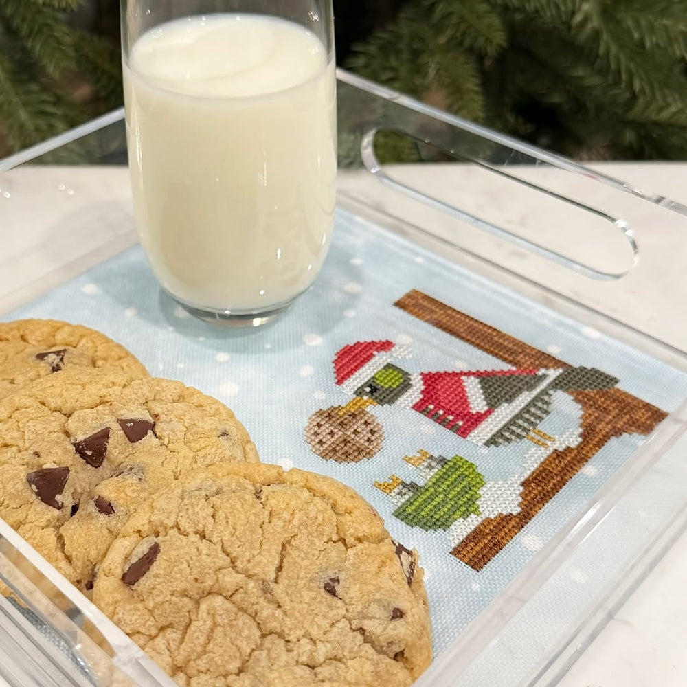 Cookies with a glass of milk on a clear tray with a Christmas-themed stitching finish underneath.