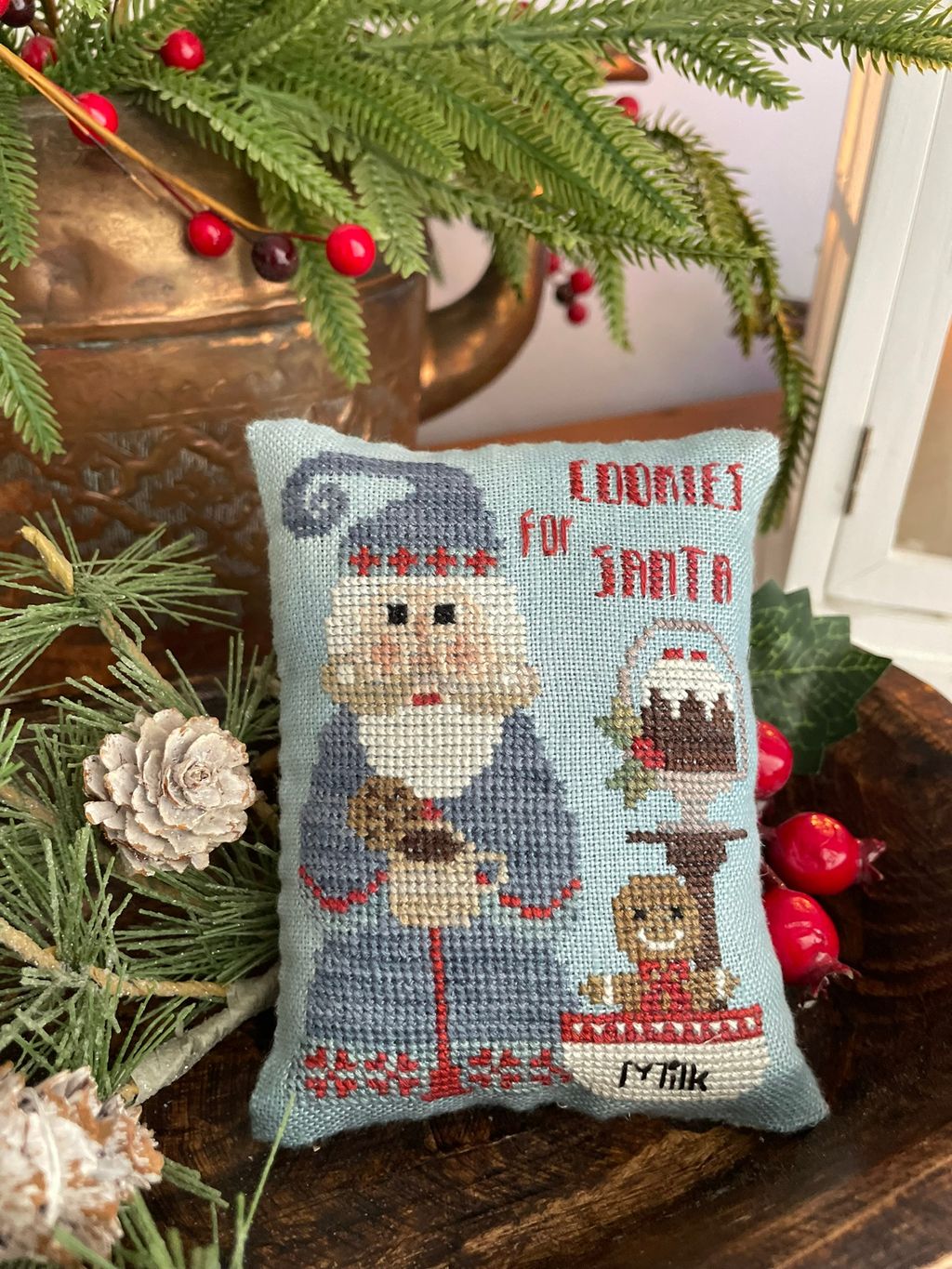 A small square cross-stitch pillow featuring Santa with cookies and the text 'COOKIES FOR SANTA,' styled on a wooden surface with pine branches, pinecones, and red berries.