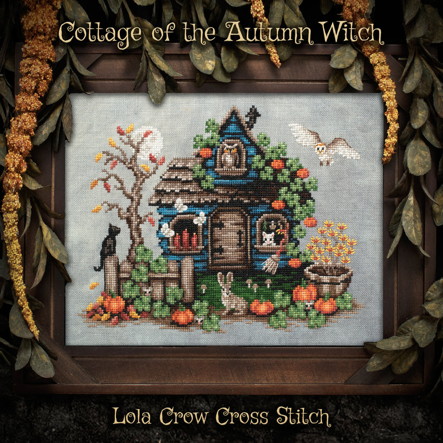 A framed cross-stitch artwork titled "Cottage of the Autumn Witch" by Lola Crow Cross Stitch, showing a small blue cottage decorated with autumn elements like pumpkins, a tree, an owl, a rabbit, and a cat, surrounded by leaves and flowers.
