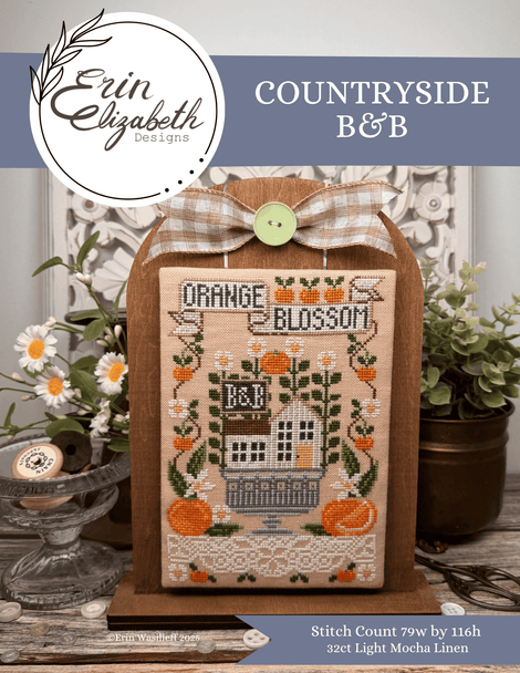 A framed cross-stitch design displayed on a wooden stand, featuring the text 'Orange Blossom' and a house with pumpkins and flowers. The image includes branding for Erin Elizabeth Designs and the title 'Countryside B&B,' with decorative flowers and plants in the background.