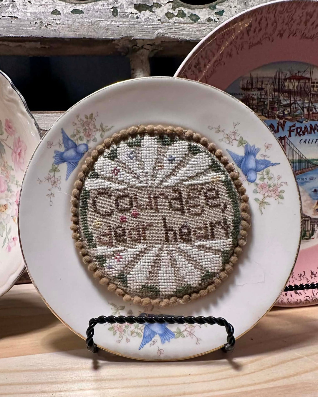 Decorative plate with a cross-stitched design and 'Courage dear heart' text, placed on a wooden surface.