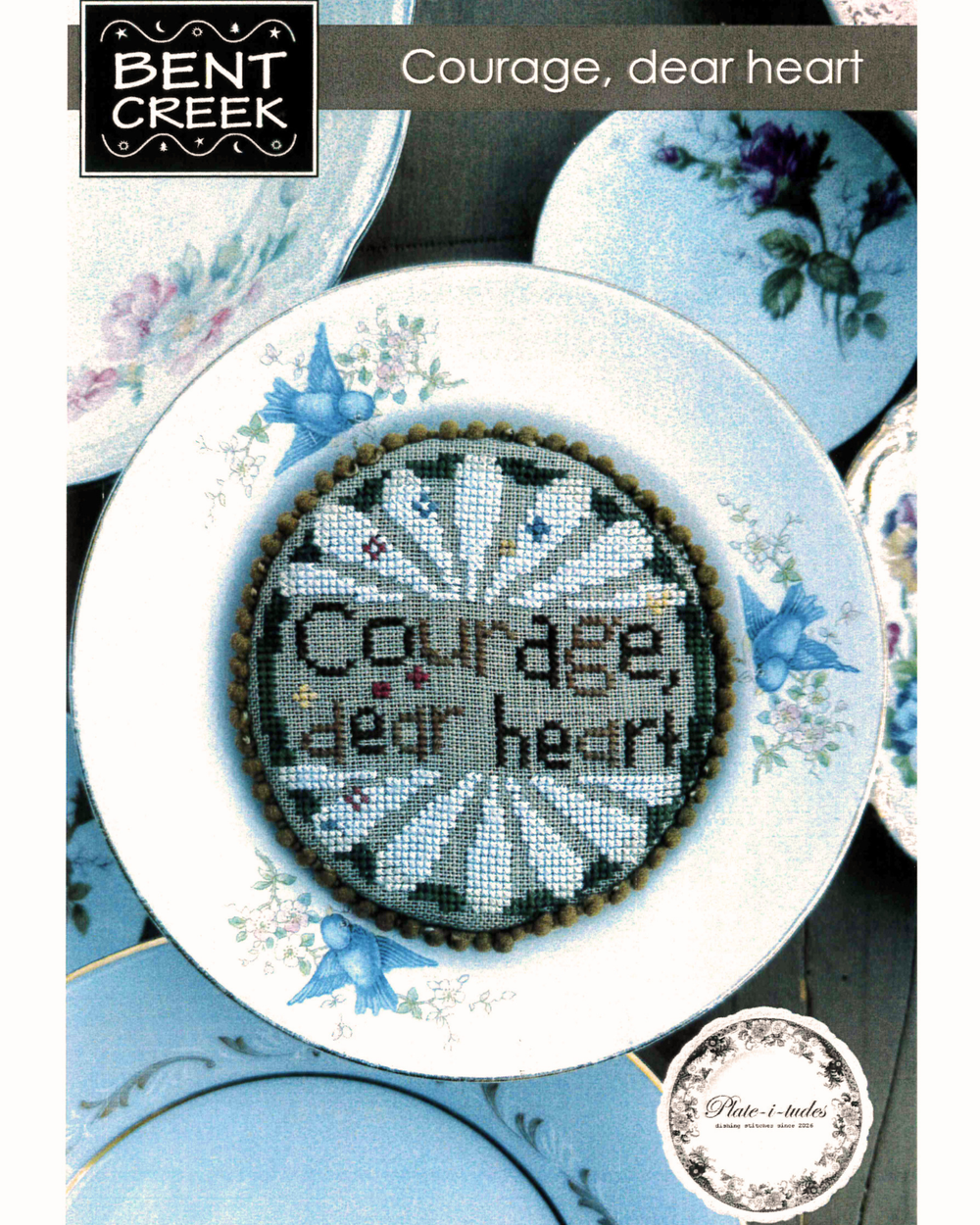 Cross-stitched design with 'Courage, dear heart' text on a decorative plate with floral patterns.
