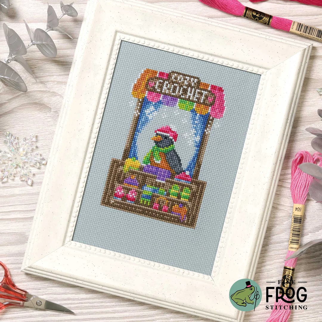 A framed cross-stitch design featuring a crocheted market stall with a frog character and the words 'CROCHET MARKET.' The white frame is photographed on a light wood surface with embroidery floss, scissors, and decorative leaves around it.