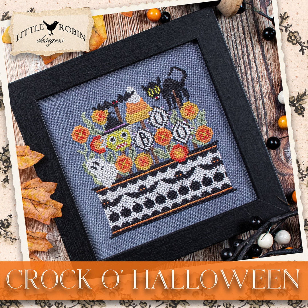 A framed cross-stitch design titled 'Crock O' Halloween' by Little Robin Designs, featuring a black cat on a witch's hat with pumpkins, ghosts, and flowers on a gray fabric background, presented in a black frame with autumn-themed decorative props.
