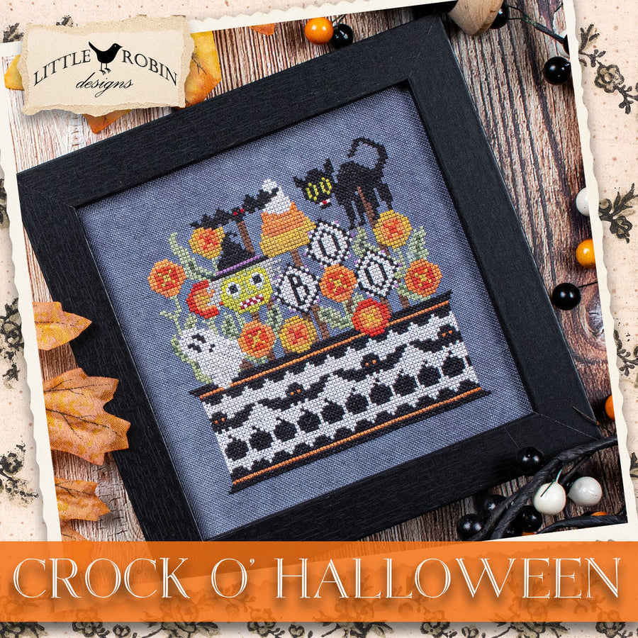 A framed cross-stitch design titled 'Crock O' Halloween' by Little Robin Designs, featuring a black cat on a witch's hat with pumpkins, ghosts, and flowers on a gray fabric background, presented in a black frame with autumn-themed decorative props.