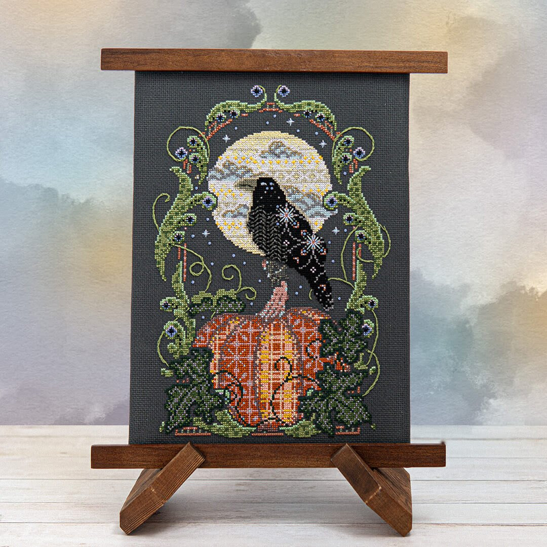 Cross-stitched piece of art with a crow and pumpkin design on a stand against a blurred background