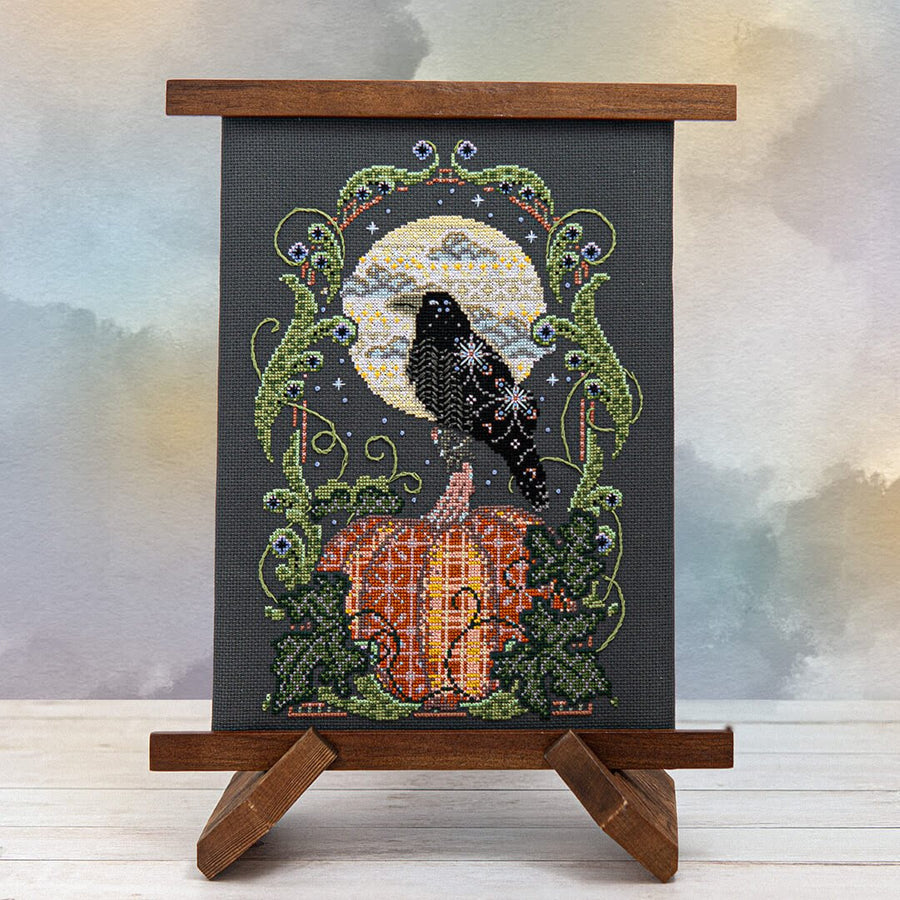 Cross-stitched piece of art with a crow and pumpkin design on a stand against a blurred background