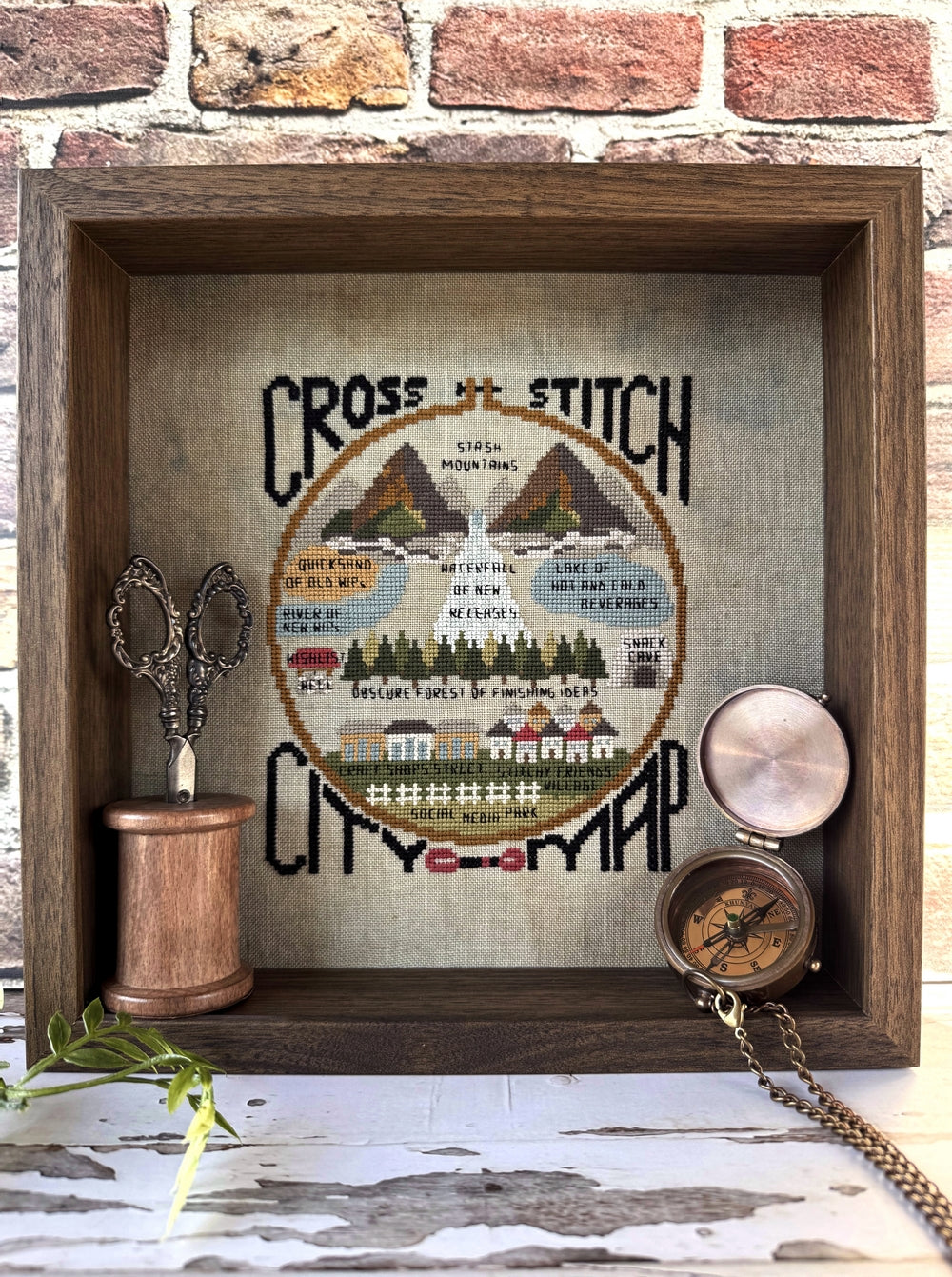 Cross-stitch city map in a wooden frame with sewing supplies on a brick wall background