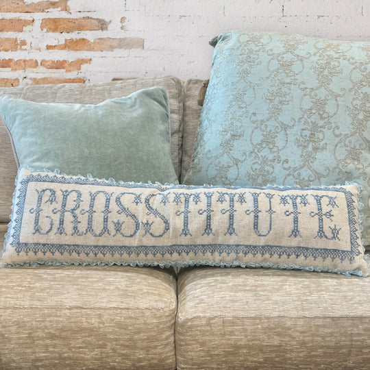Decorative pillow with 'CROSSSTITUTE' embroidery on a couch against a brick wall.