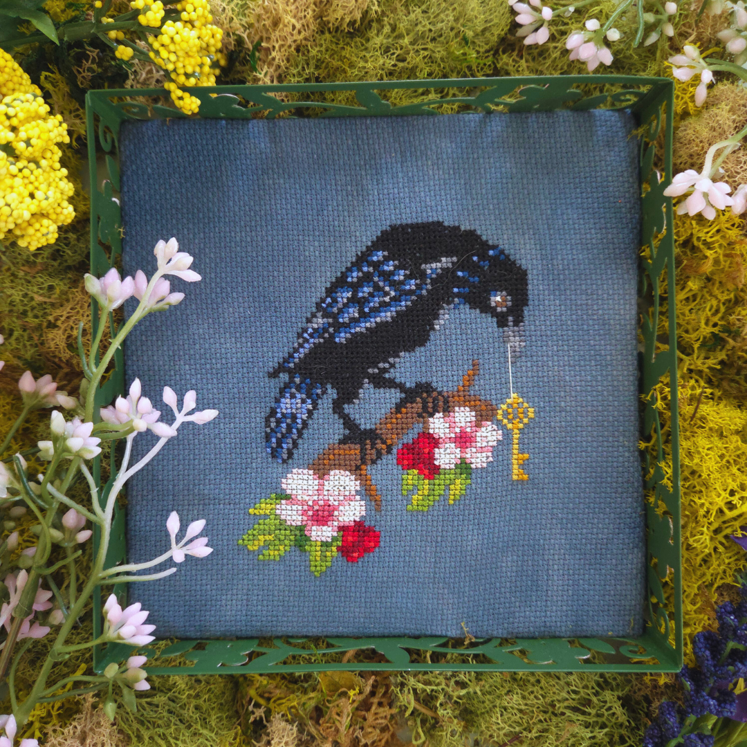 Embroidery of a blue bird on a branch with flowers, framed by natural elements.
