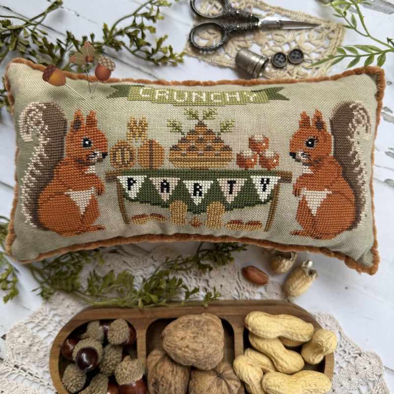 A rectangular embroidered cushion with a autumn squirrel party design, styled on a white lace surface with nuts, scissors, and dried greenery.