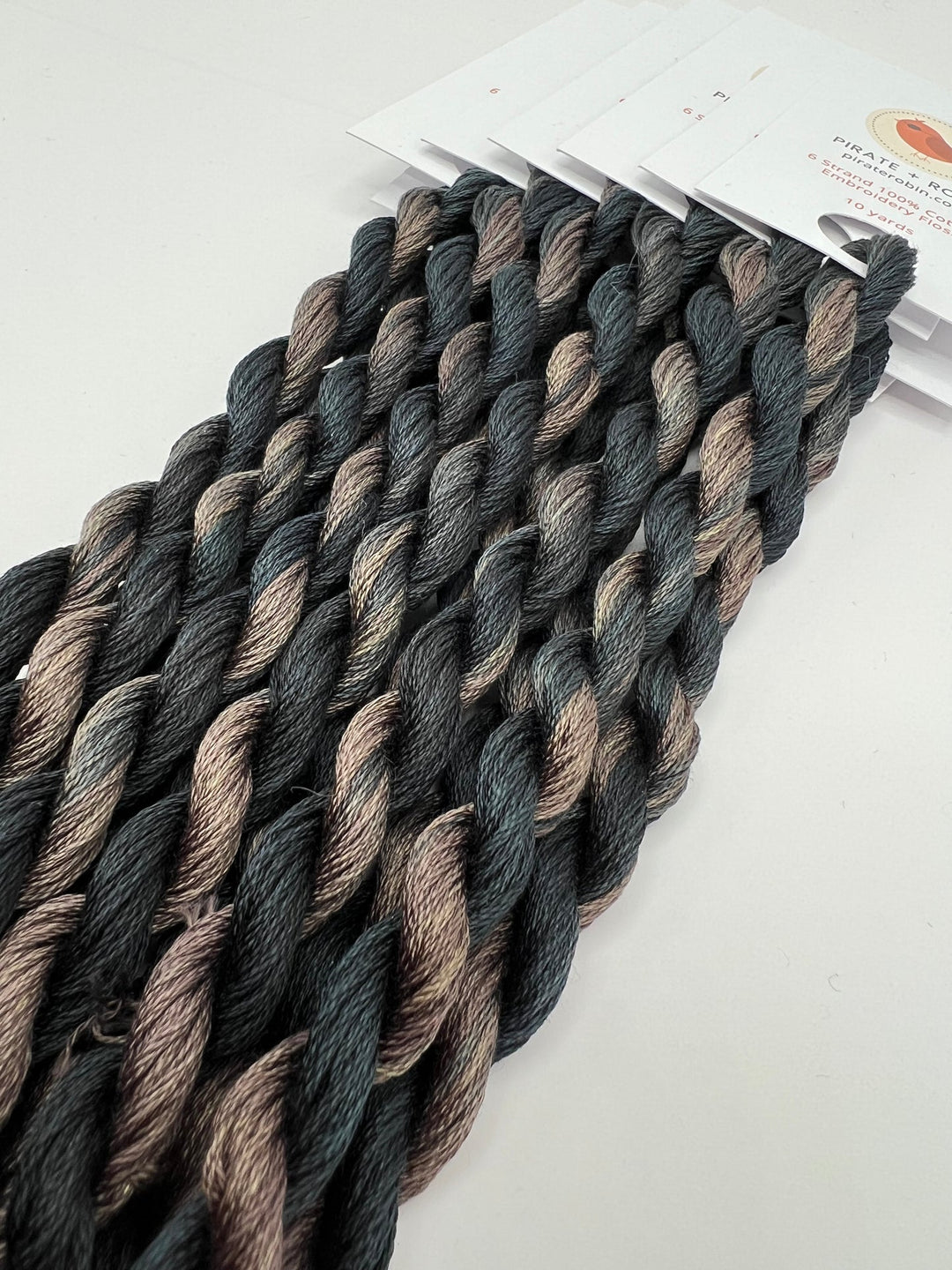Braided thread skeins in a variegated black, gray, and brown colorway, shown with a white paper header card on a plain white background.