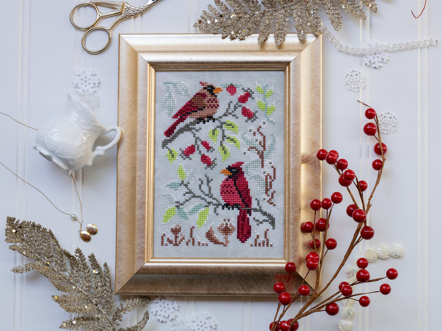 Framed cross-stitched picture of cardinals on a branch with red berries, surrounded by decorative items.