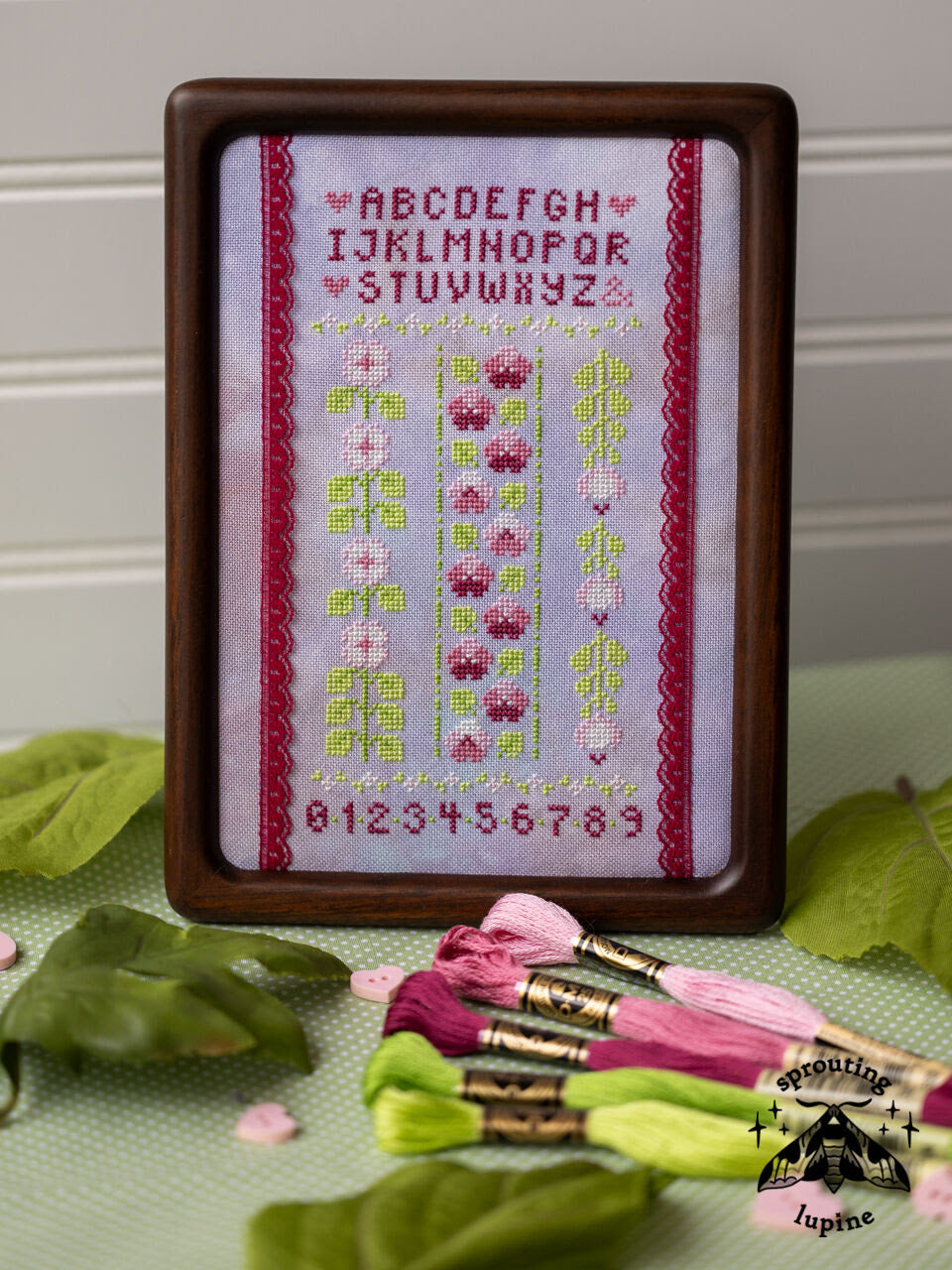 Framed cross-stitched sampler with floral patterns and letters, surrounded by embroidery threads on a green surface.