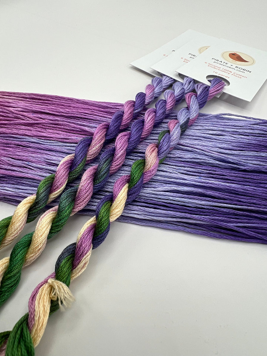 Multicolor embroidery floss yarn skeins in shades of purple, green, and cream, with three twisted braids in the foreground and a product tag attached at the top.